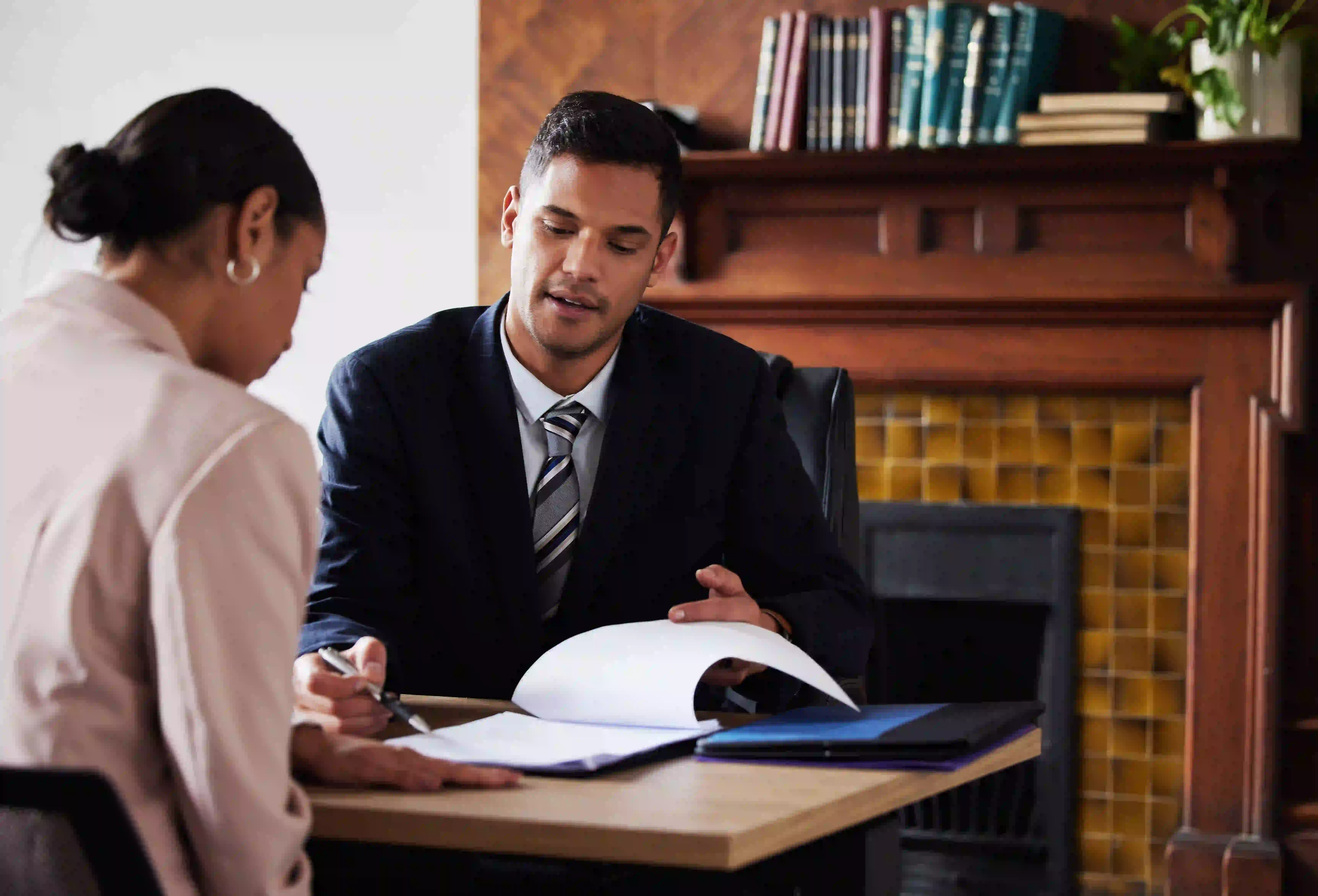 A professional male attorney in a dark suit and striped tie sits at a wooden desk in a classic office, reviewing legal documents with a female client.